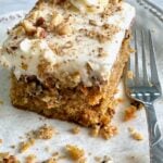 slice of carrot cake with icing and nuts on a white plate with a fork beside it