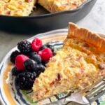 a slice of meat lover's quiche with berries on a plate and skillet in the background.