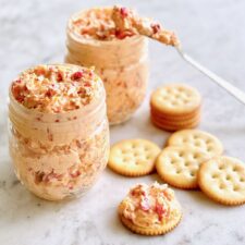 pimento cheese in mason jars with crackers and a spreader dipping in the cheese.