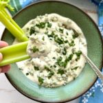fingers dipping celery stick into homemade blue cheese dip in a green bowl.
