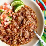 Mexican Shredded Beef in a white bowl with limes and pico de Gallo on the side.