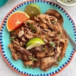 plate of carnitas made in a dutch oven and served with lime and orange slices.