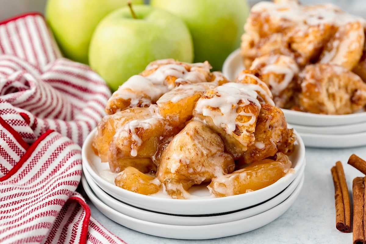 A plate of glazed apple fritters is placed in the foreground with a red and white striped cloth to the side. In the background, there are fresh green apples and another plate of fritters. Two cinnamon sticks are also visible on the right.