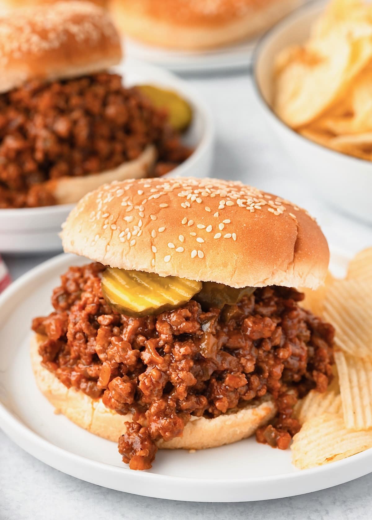 This image shows a sloppy Joe topped with pickles served on a white plate with potato chips.