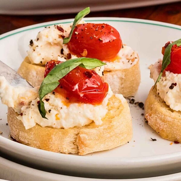 Ricotta Cheese Dip being spread on a crostini topped with tomatoes and basil.
