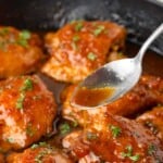 A spoon drizzles sauce over glazed chicken pieces in a skillet, garnished with chopped herbs.