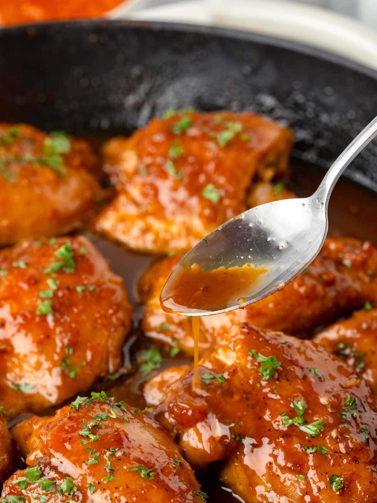 A spoon drizzles sauce over glazed chicken pieces in a skillet, garnished with chopped herbs.