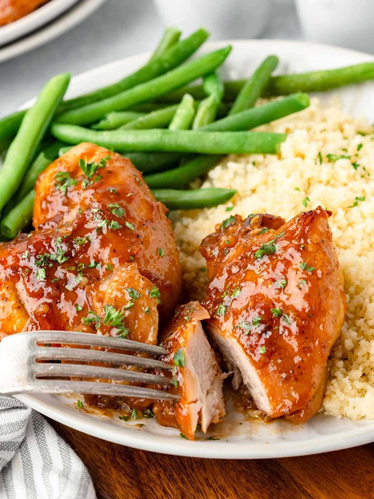 A plate with glazed chicken breast, green beans, and couscous. A fork is cutting into the chicken.