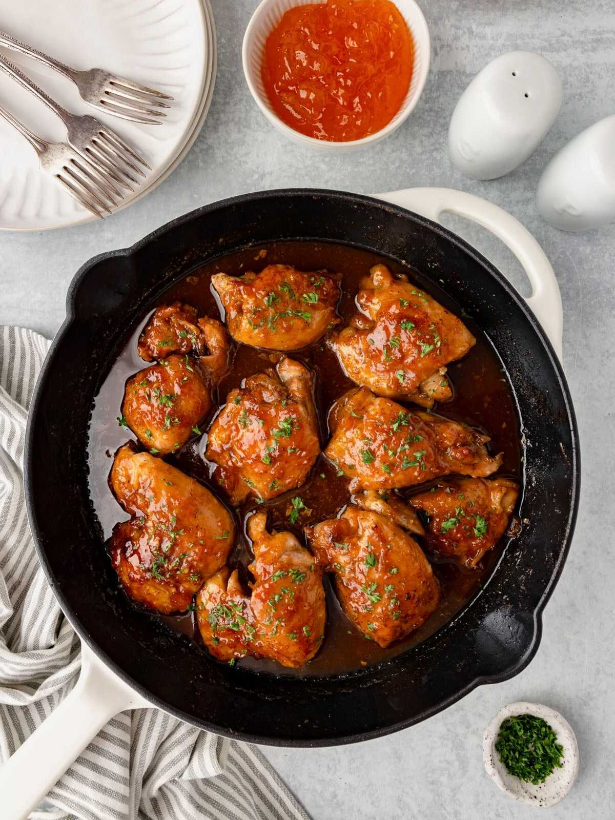 Chicken thighs glazed with sauce in a cast iron skillet, garnished with herbs, with plates and sauce nearby.