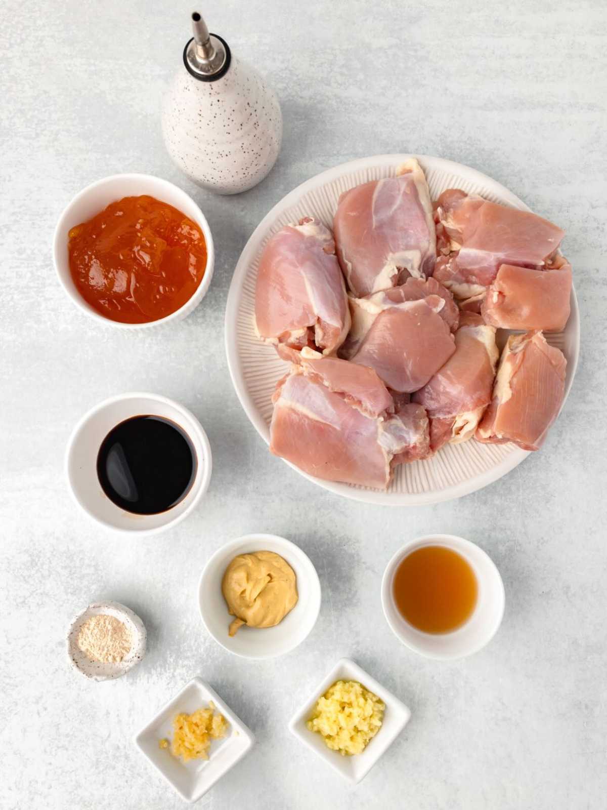 Raw chicken pieces on a plate surrounded by bowls of sauces, seasonings, and a bottle of oil on a light surface.