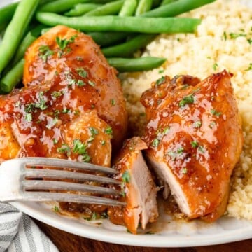 Fork cutting into saucy chicken breast with green beans and couscous on a plate.