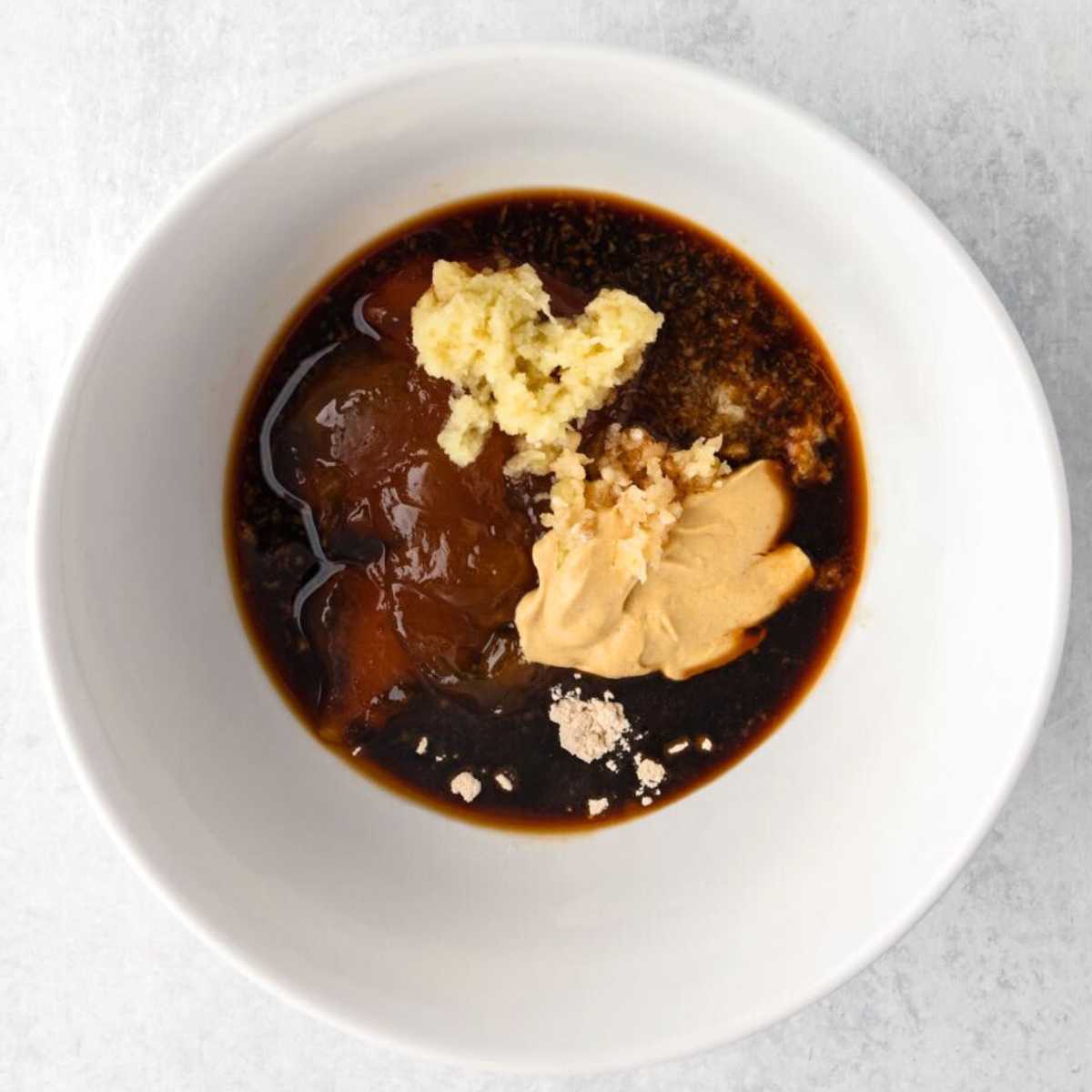 A white bowl with soy sauce, minced ginger, peanut butter, and brown paste ingredients on a light surface.