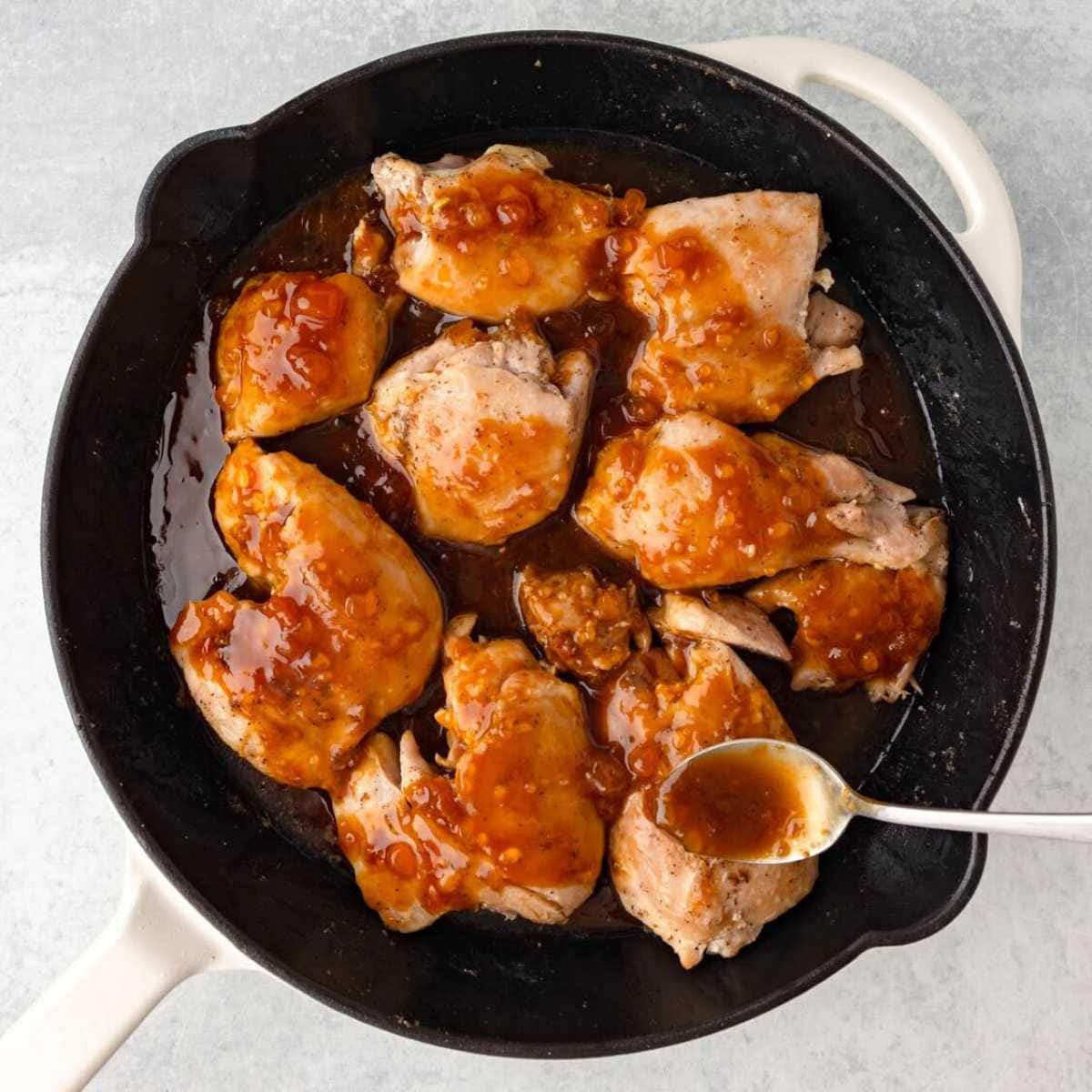 Chicken thighs covered in brown sauce cooking in a black skillet, with a spoon scooping up the sauce.