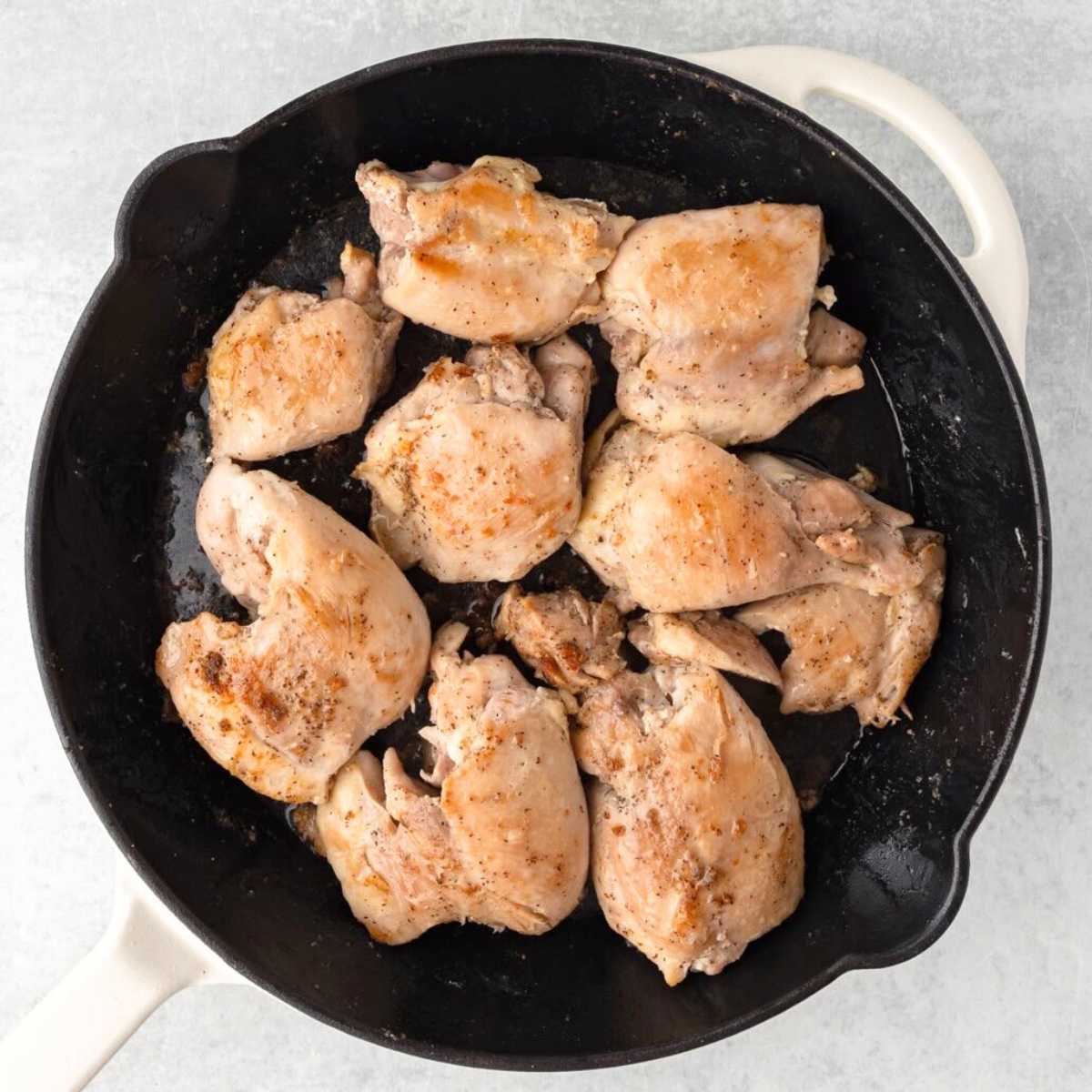 Cooked chicken pieces in a black skillet on a light gray surface, seasoned with spices.