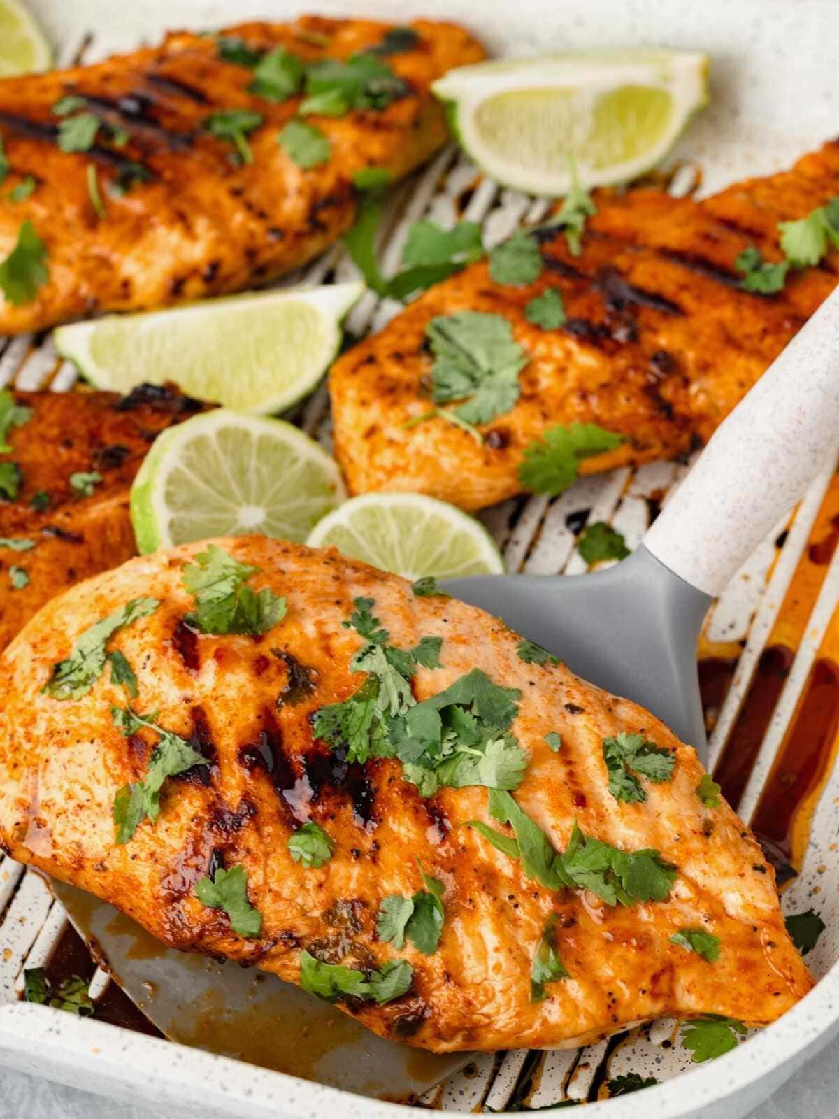 Grilled chicken breasts garnished with cilantro and lime slices on a grill pan with a spatula.