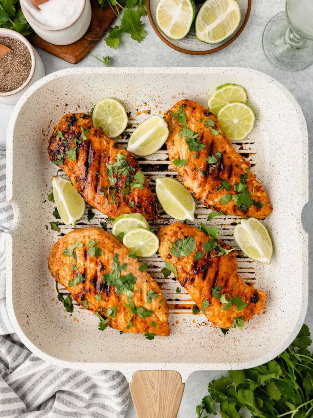 Grilled chicken breasts in a grill pan, garnished with cilantro and lime wedges, surrounded by more lime slices.