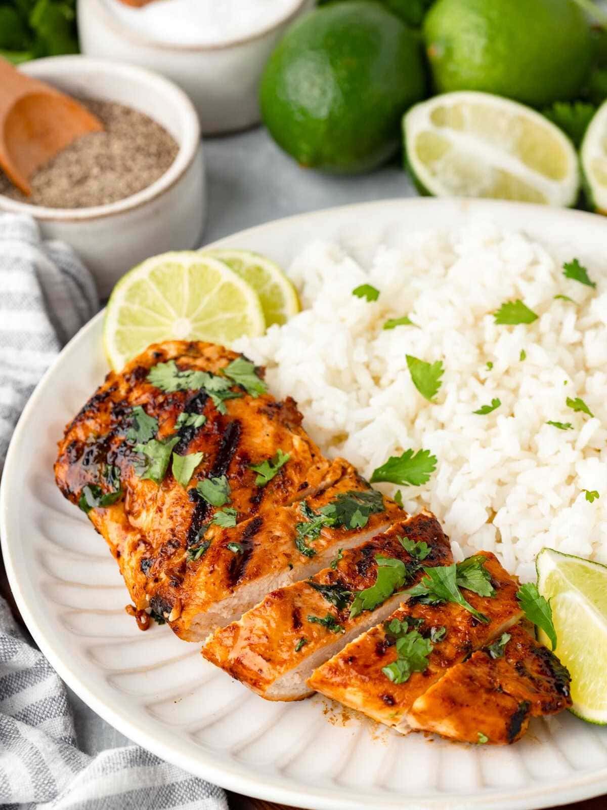 Sliced grilled chicken with cilantro, lime, and white rice on a plate, with limes and seasonings in the background.
