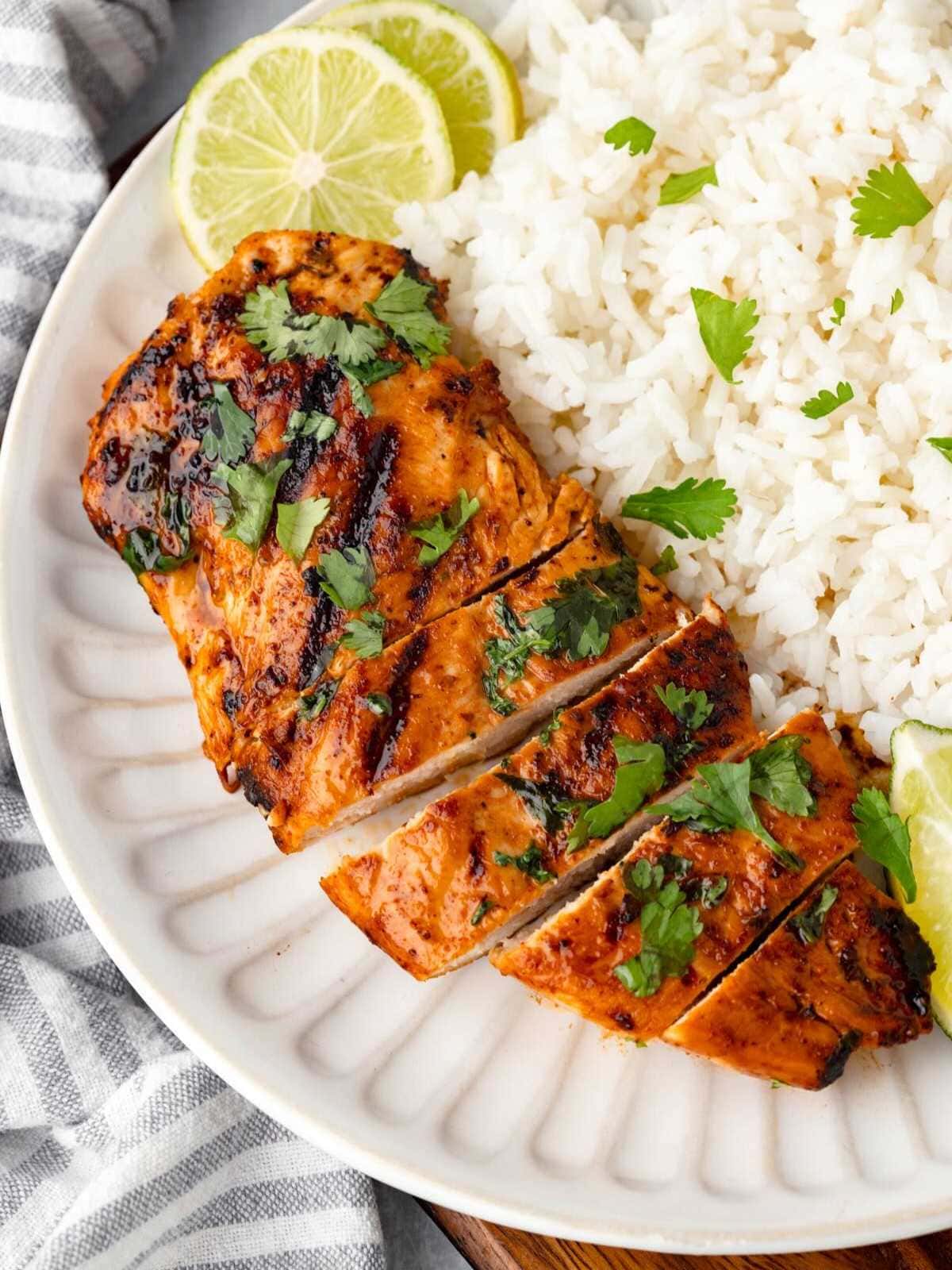 Grilled chicken with cilantro, sliced next to white rice and lime wedges on a white plate.