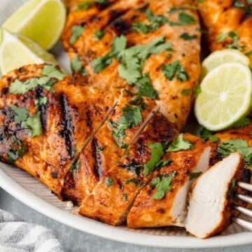 Grilled chicken breast sliced and garnished with cilantro, served with lime wedges on a plate.