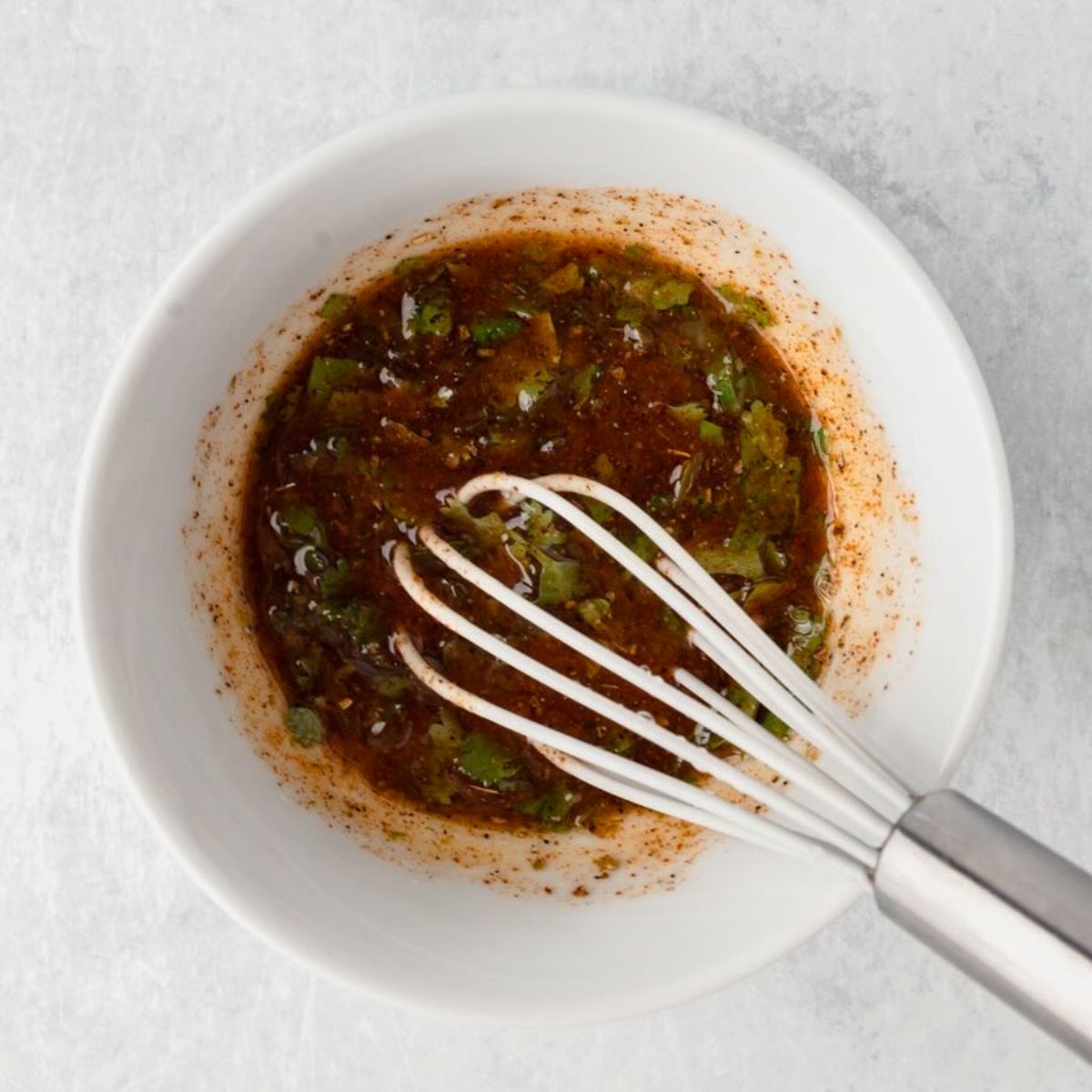 A whisk in a white bowl with cilantro marinade mixed with herbs.