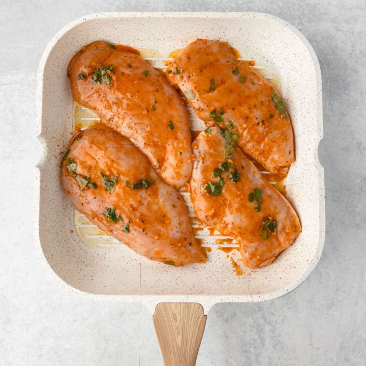 Raw, marinated chicken breasts in a white grill pan with a wooden handle, ready to be cooked.