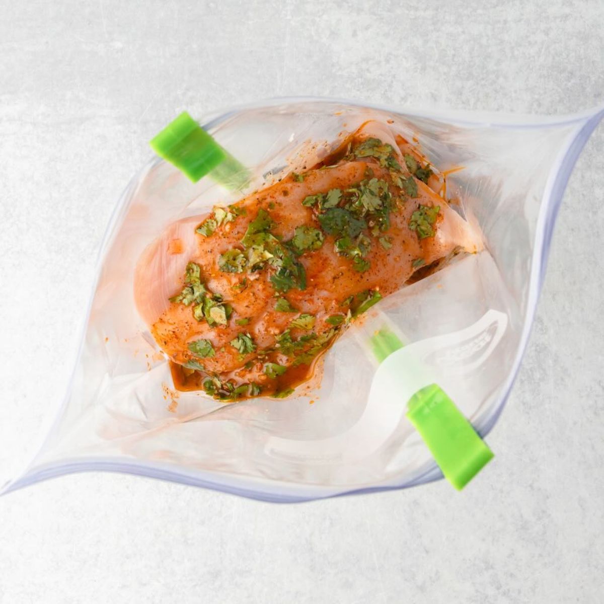 Raw chicken breast marinating in herbs and spices inside a resealable plastic bag on a light surface.