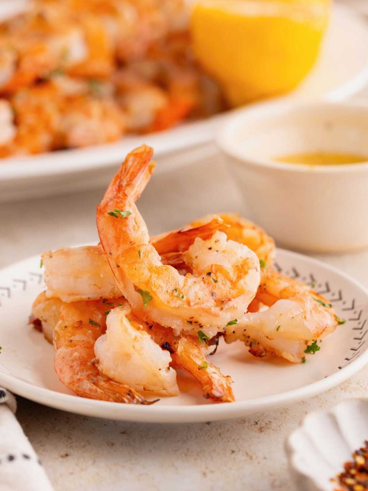 Cooked shrimp garnished with herbs on a white plate, served with a lemon wedge and dipping sauce.