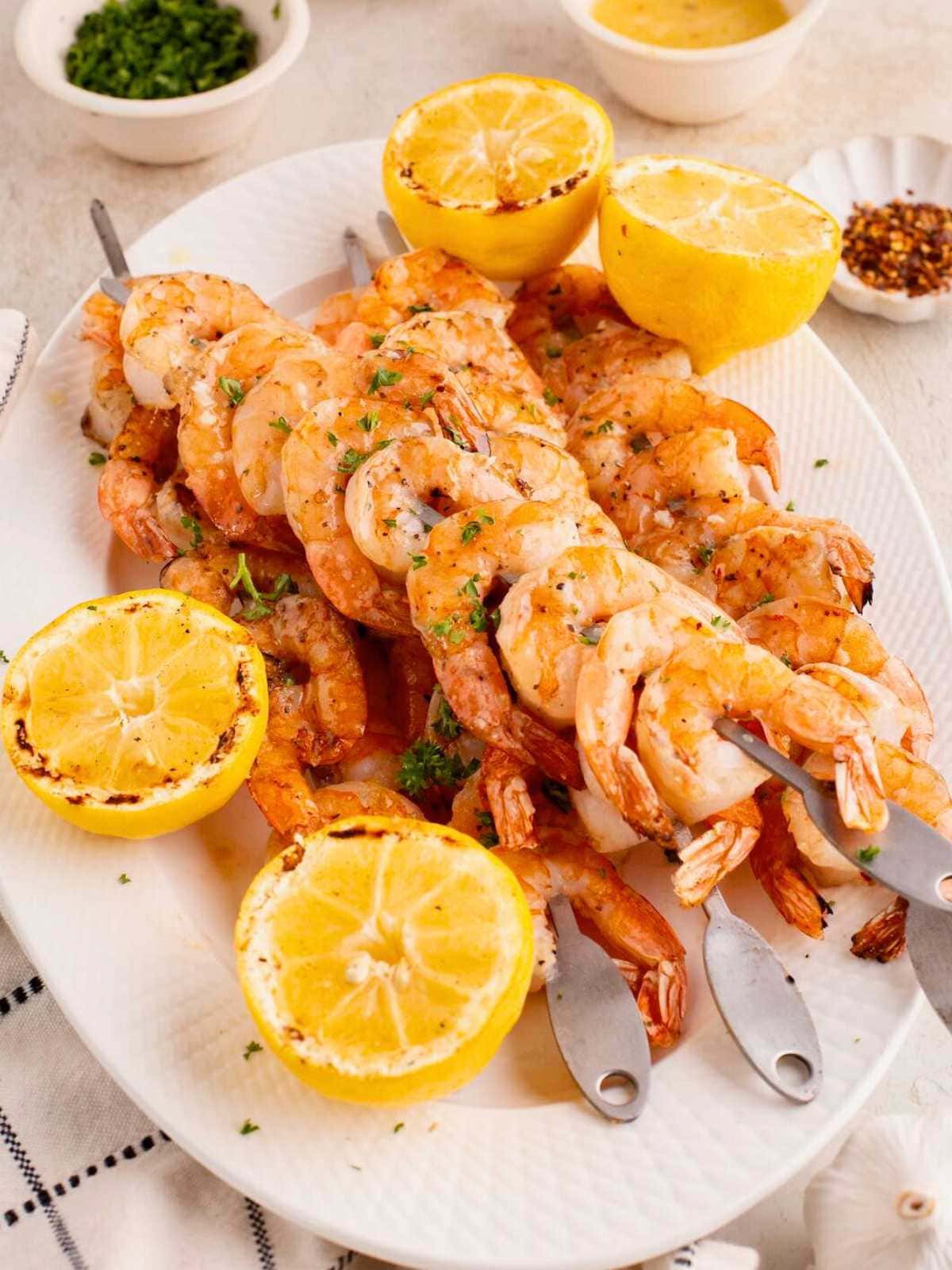 Grilled shrimp skewers on a white plate with grilled lemon halves and sprinkled herbs.