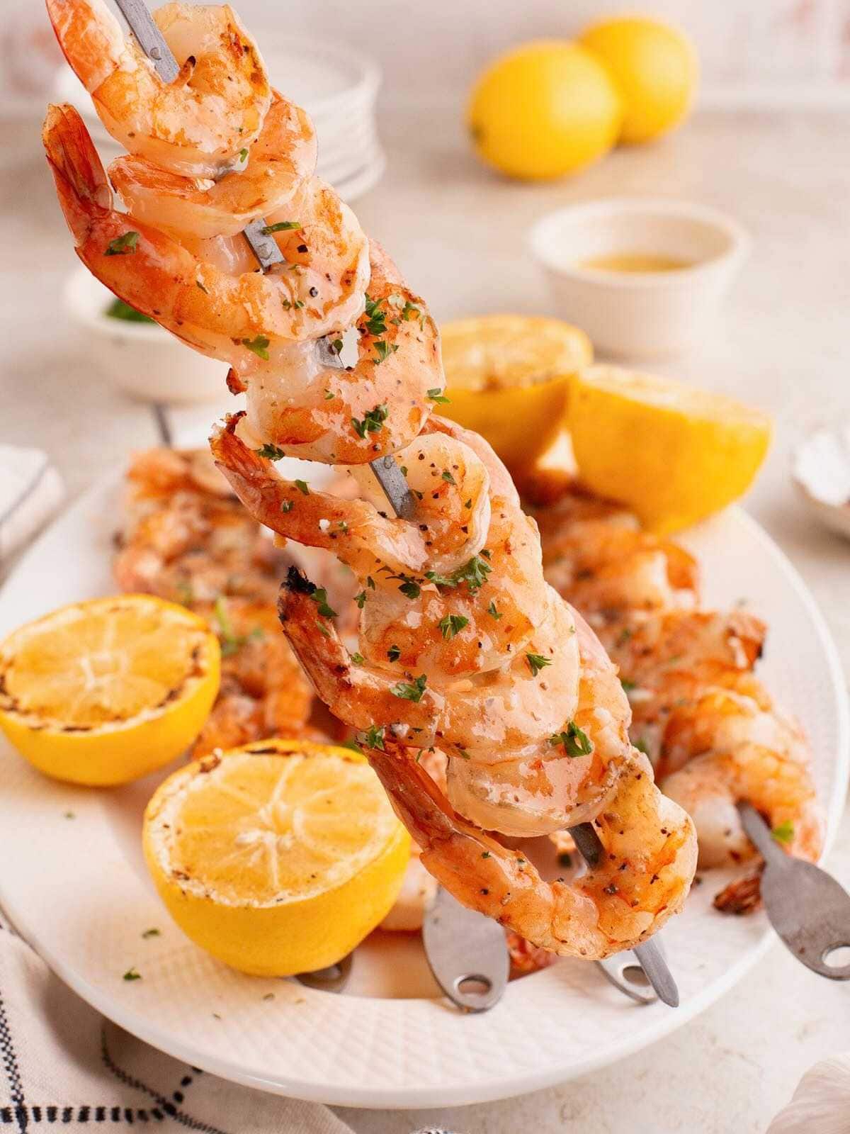 Grilled shrimp skewers garnished with herbs, served with halved lemons on a white plate.