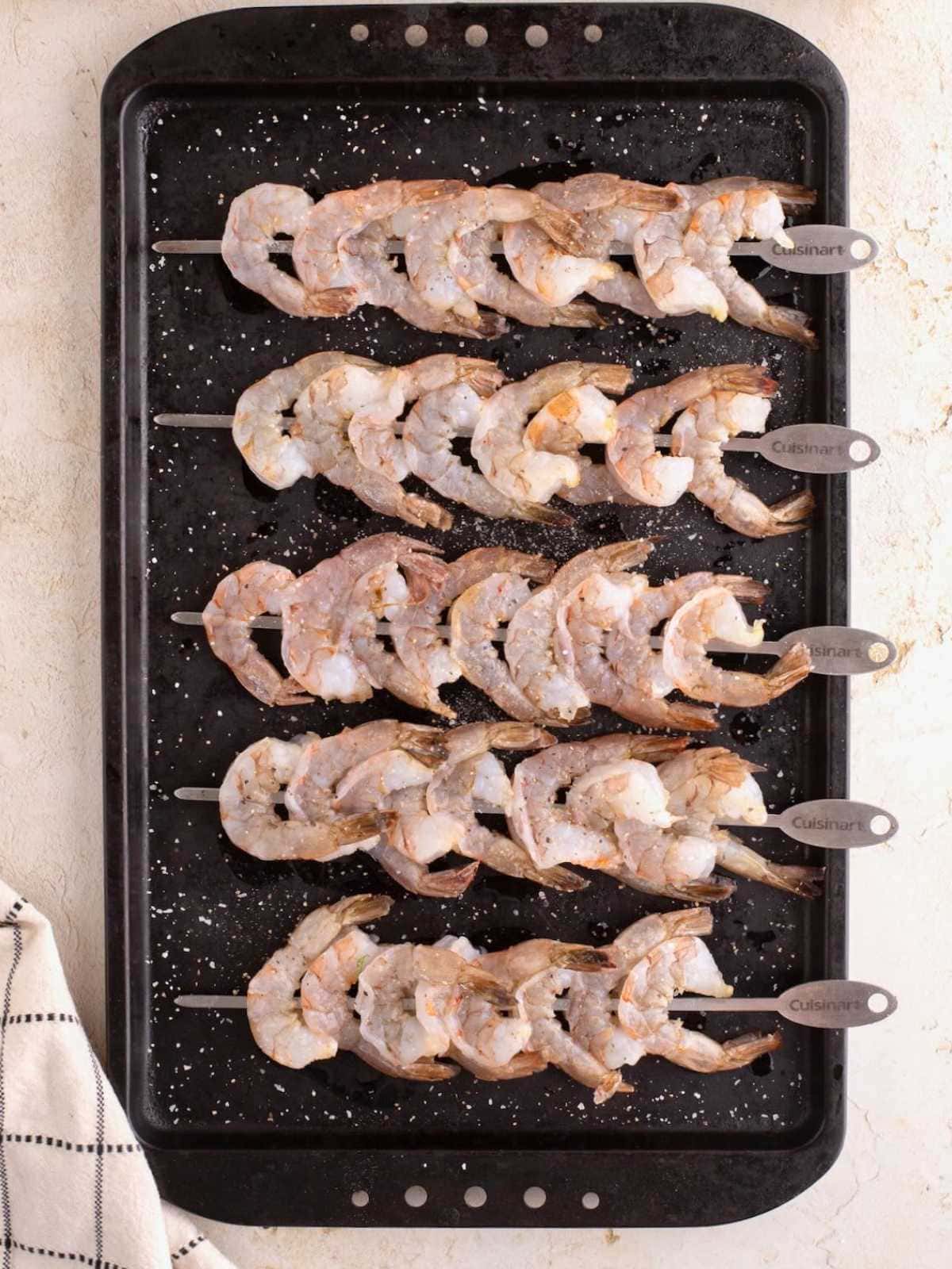 Raw shrimp skewers arranged on a black baking sheet, sprinkled with salt, ready for cooking.