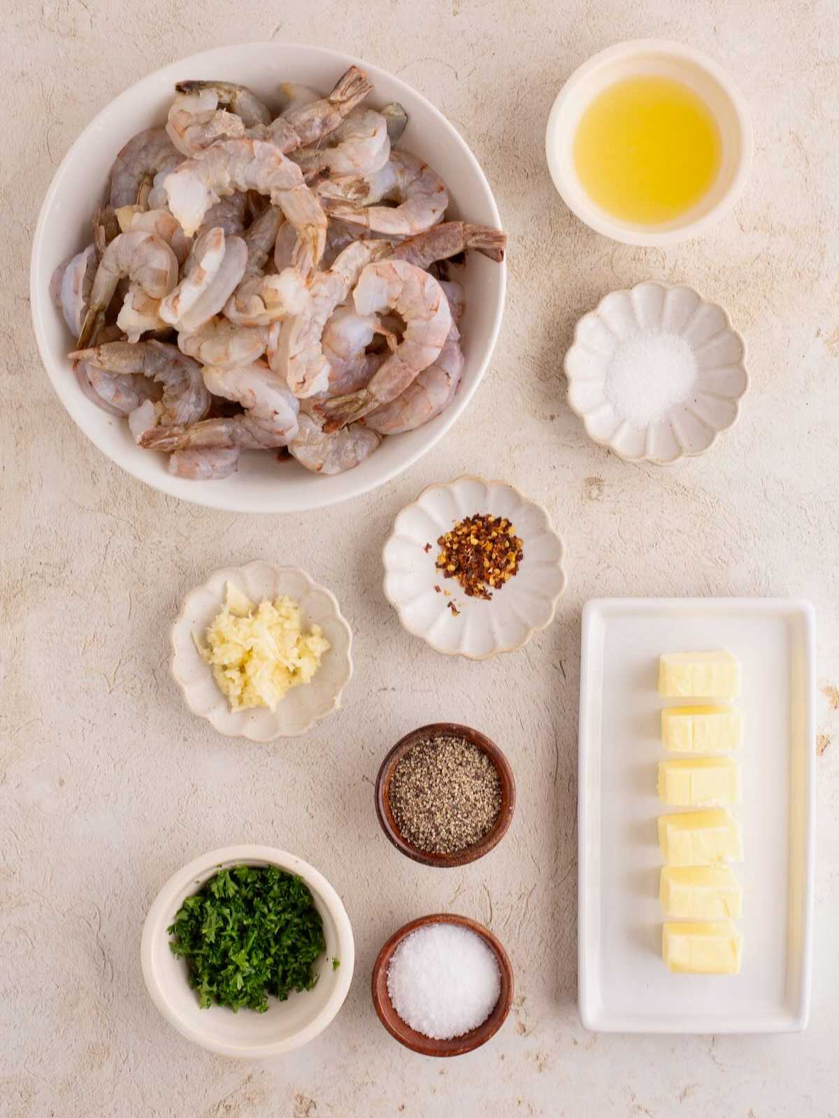 Raw shrimp, butter cubes, chopped parsley, minced garlic, spices, salt, pepper, and lemon juice in separate bowls.