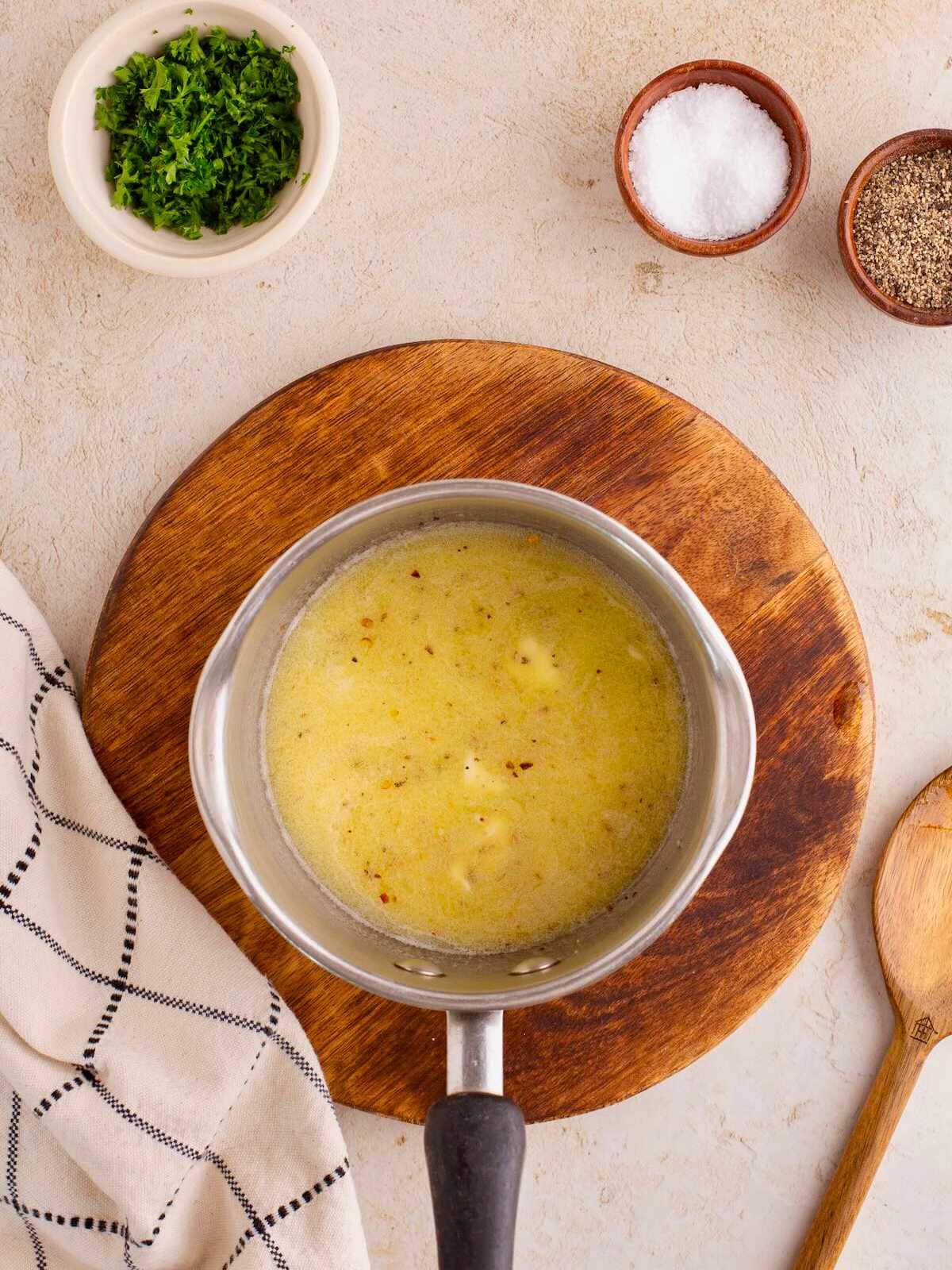 A saucepan with melted butter on a wooden board, surrounded by herbs, salt, pepper, a towel, and a wooden spoon.