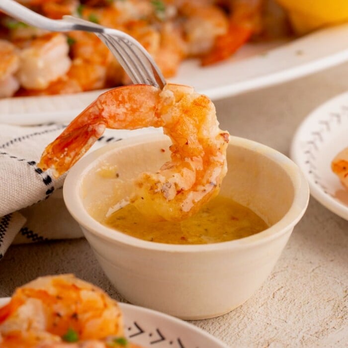 A fork dips a grilled shrimp into a small bowl of melted garlic butter sauce.