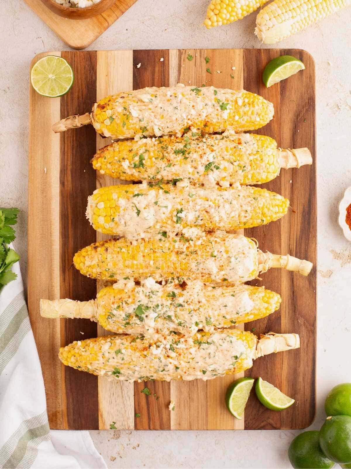 Six ears of Mexican street corn with creamy sauce and herbs on a wooden board, garnished with lime wedges.