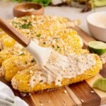 A brush spreads creamy sauce on grilled corn topped with cheese and herbs, with lime wedges on the side.