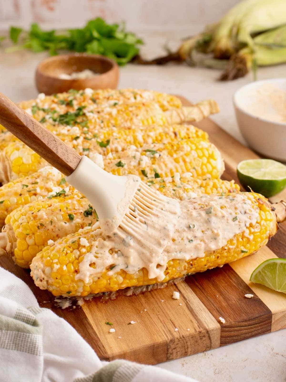 A brush spreads creamy sauce on grilled corn topped with cheese and herbs, with lime wedges on the side.