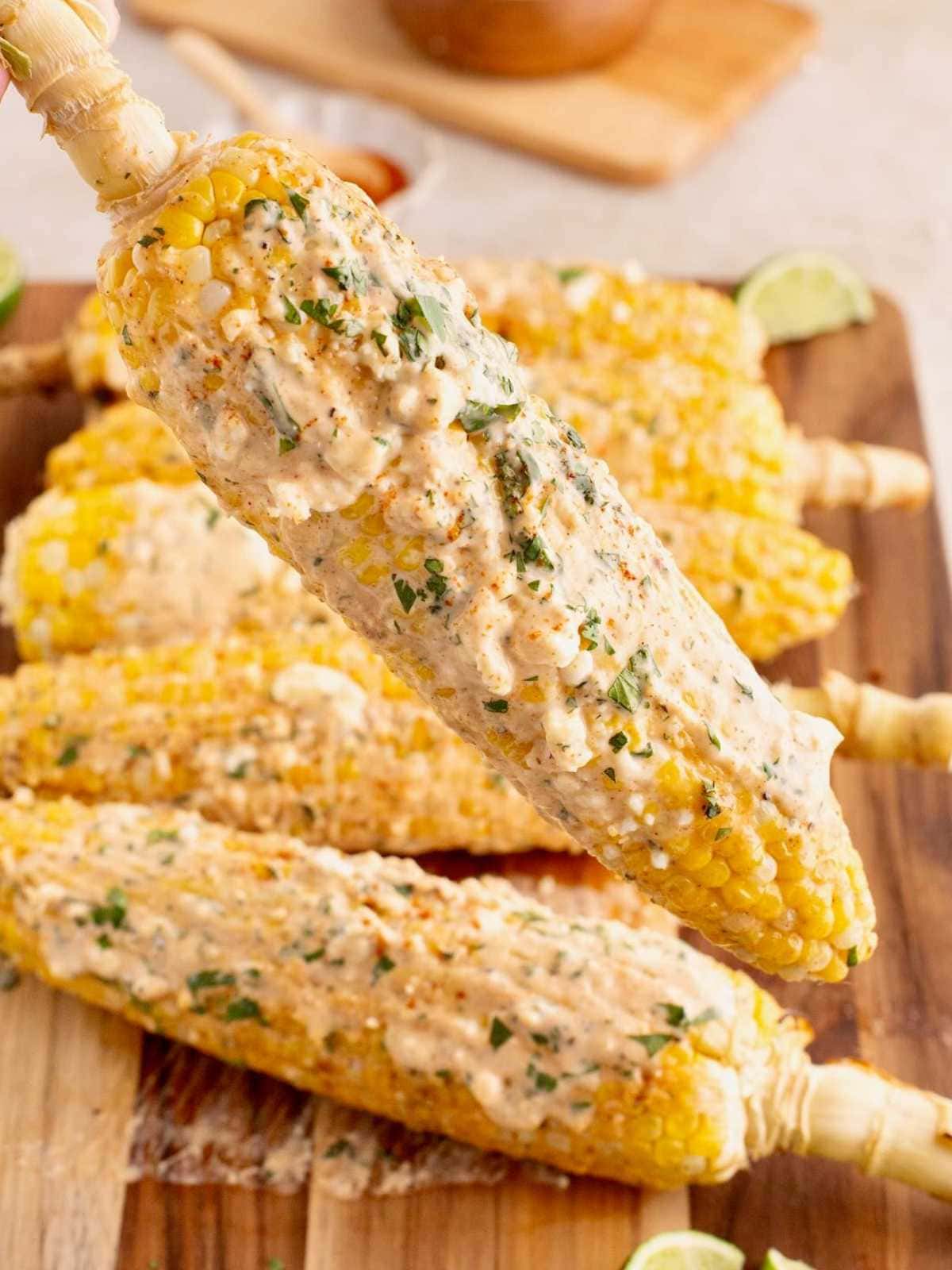 Elote (Mexican street corn) covered in creamy sauce, cheese, and herbs, served on a wooden board with limes.