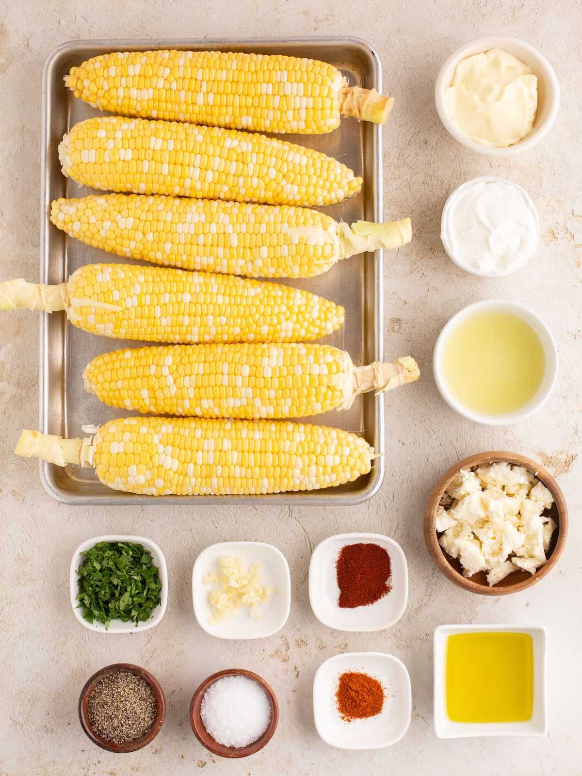 Six ears of corn on a tray, surrounded by bowls of spices, herbs, cheese, oil, sauce, and mayonnaise.