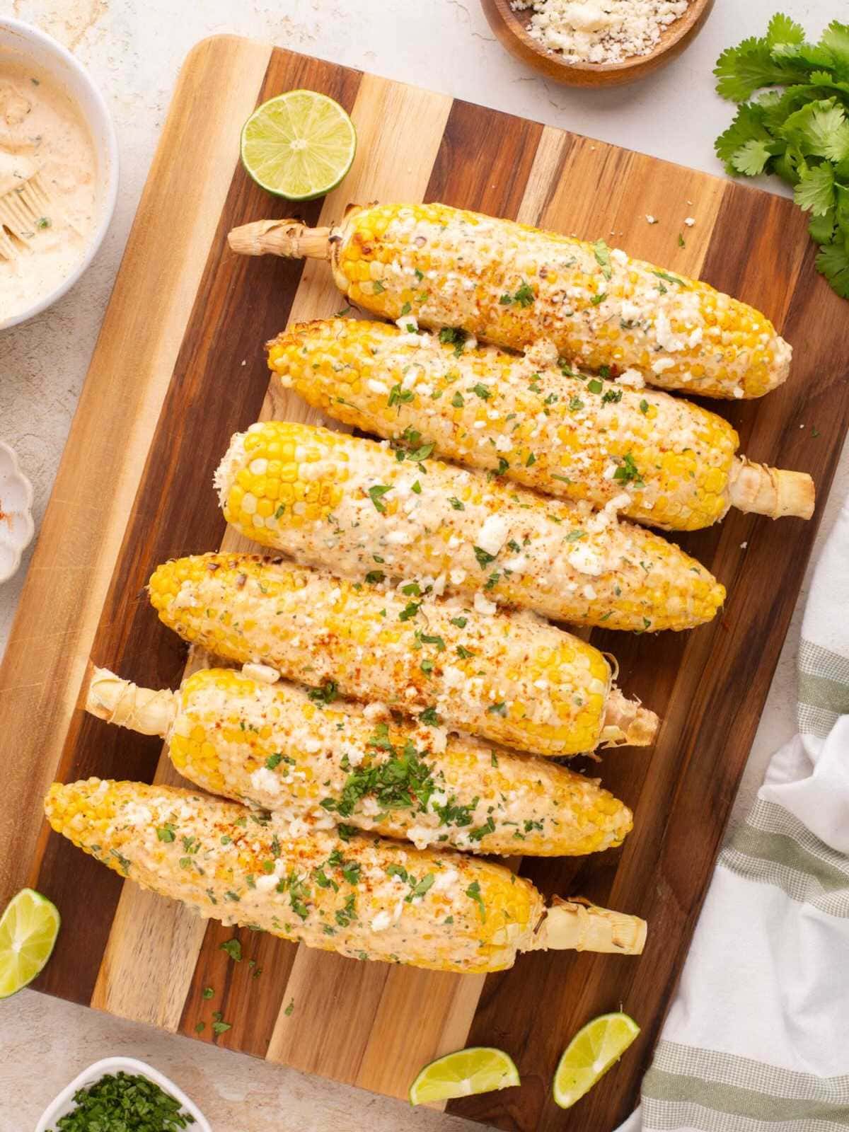 Six Mexican street corn cobs on a wooden board, topped with cheese, herbs, and lime wedges around.