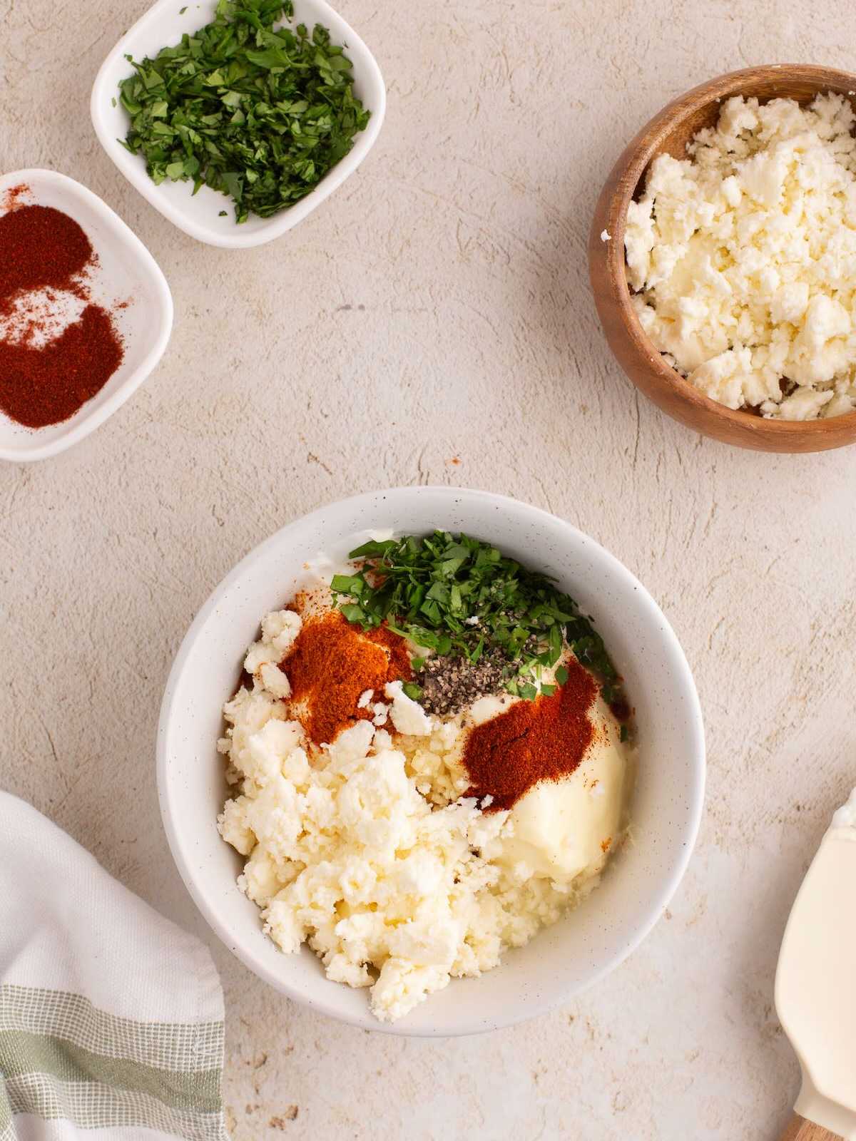 A bowl with cheese, paprika, pepper, and chopped herbs, surrounded by small bowls of spices and herbs.