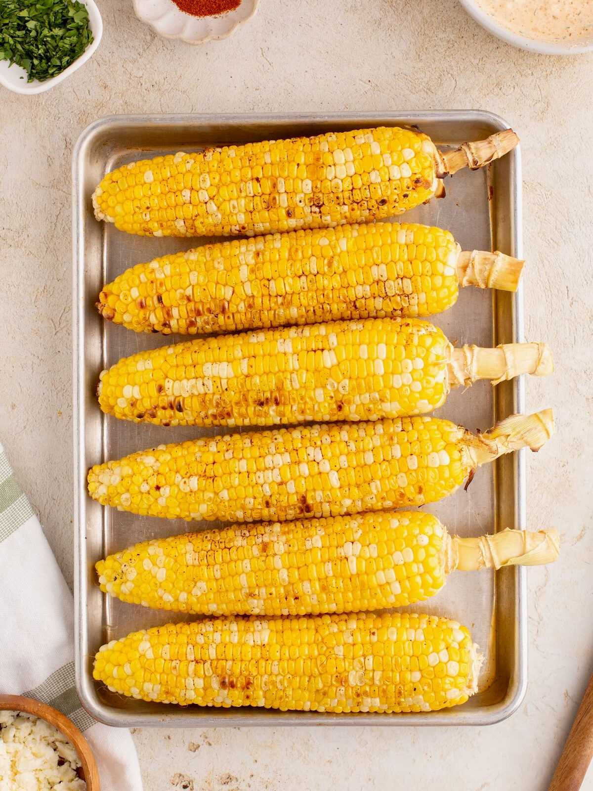 Six grilled corn cobs lined up on a metal tray, with husks removed, on a light surface.