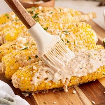 A brush spreads creamy sauce over grilled Mexican street corn, topped with herbs and cheese.