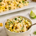 A bowl of creamy corn pasta salad with herbs, with a fork and lime wedge nearby on a marble surface.