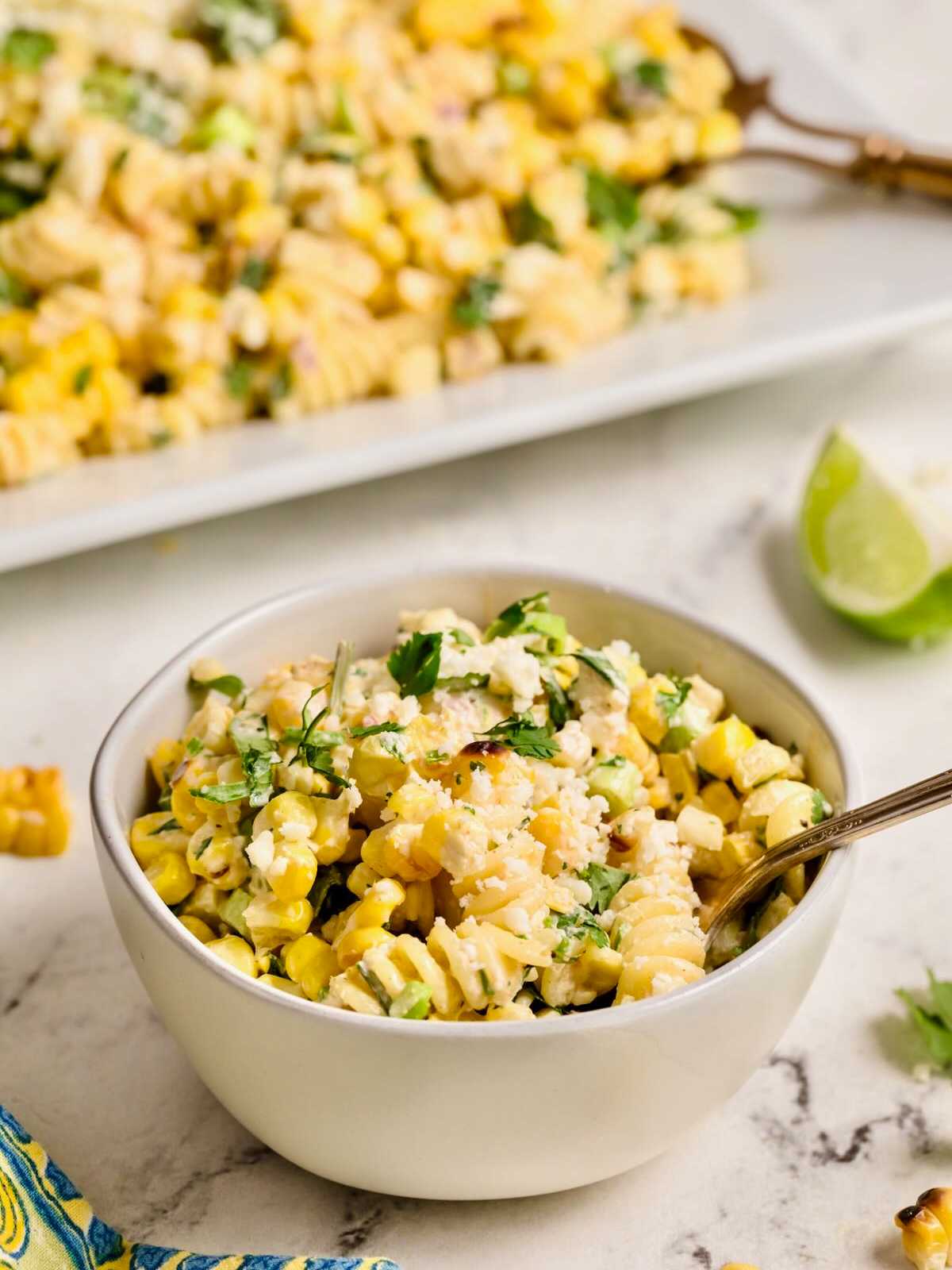 A bowl of creamy corn pasta salad with herbs, with a fork and lime wedge nearby on a marble surface.