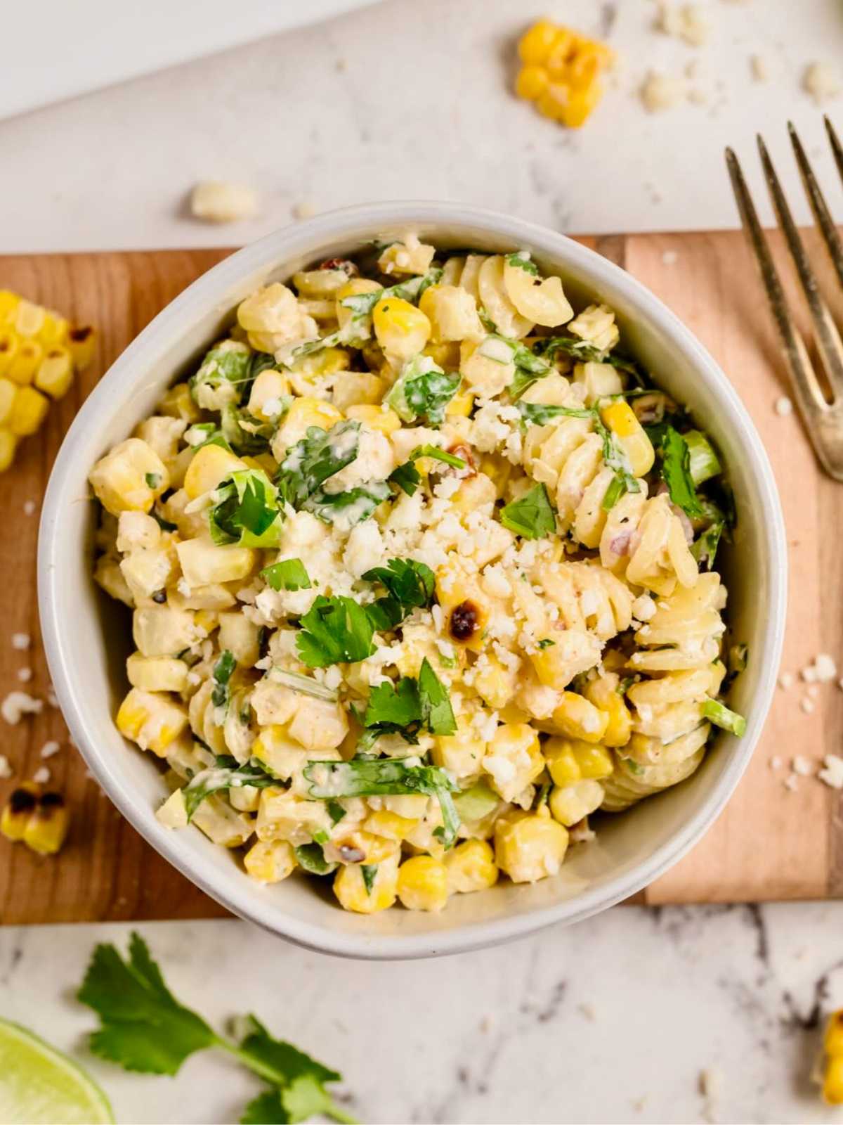 A bowl of creamy Mexican street corn salad topped with cilantro and cheese sits on a wooden board.