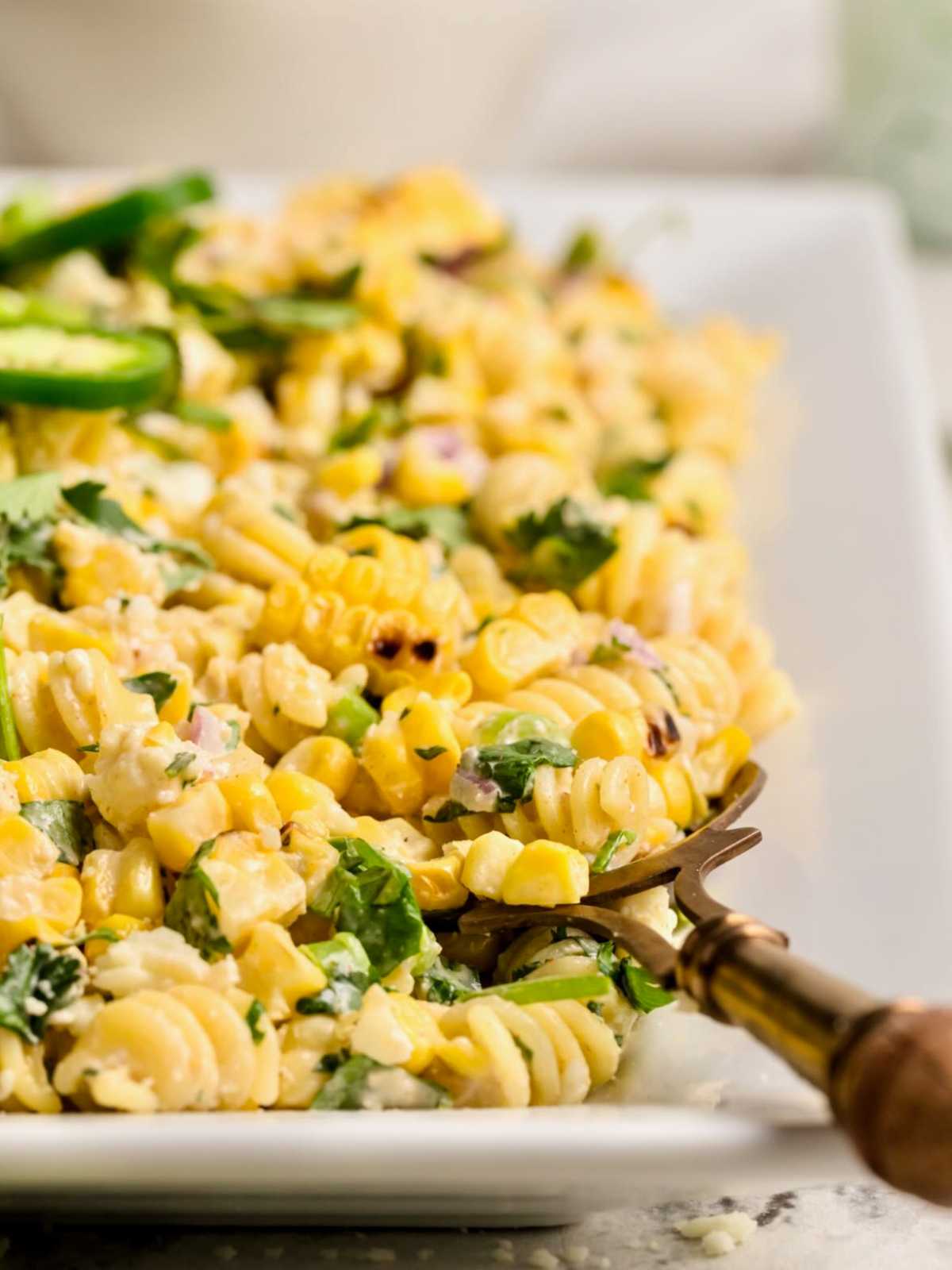A close-up of creamy pasta salad with corn, herbs, and cheese on a white plate with a serving fork.