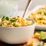 A white bowl of Mexican street corn salad garnished with cilantro, with a spoon and lime wedge nearby.