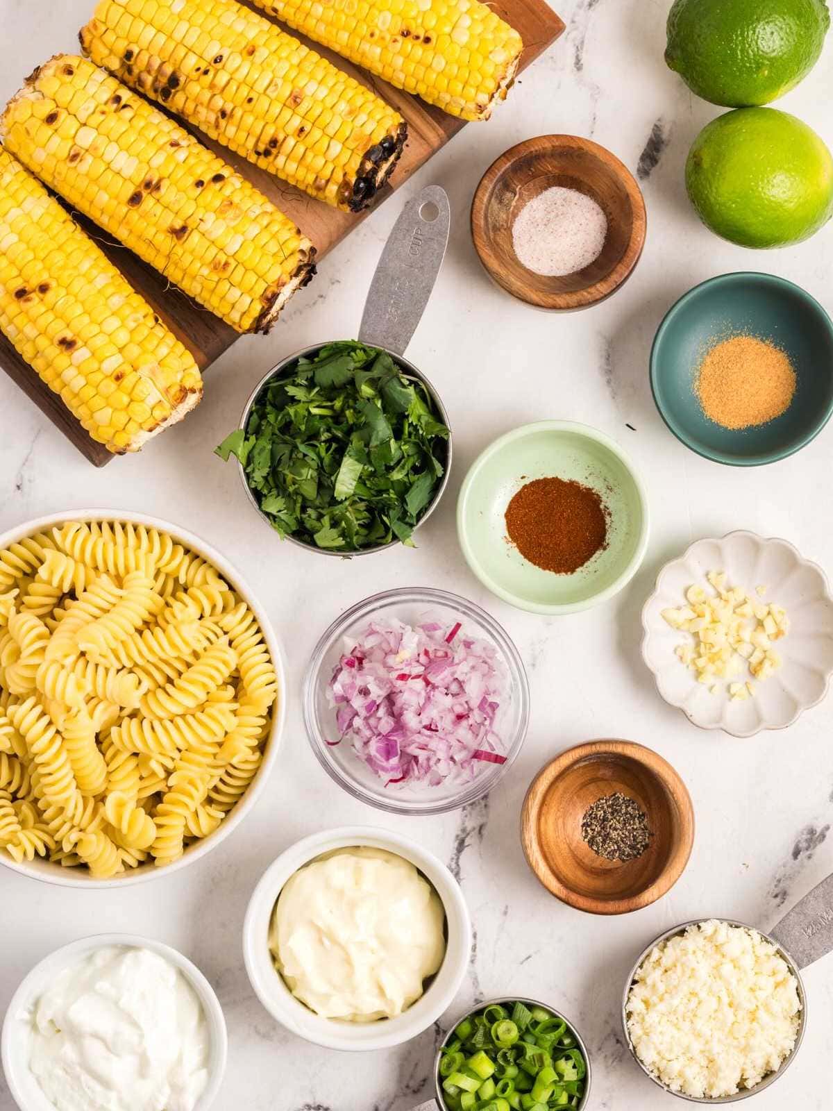 Cooked corn, rotini pasta, herbs, seasonings, mayonnaise, cheese, and lime ingredients on a white surface.