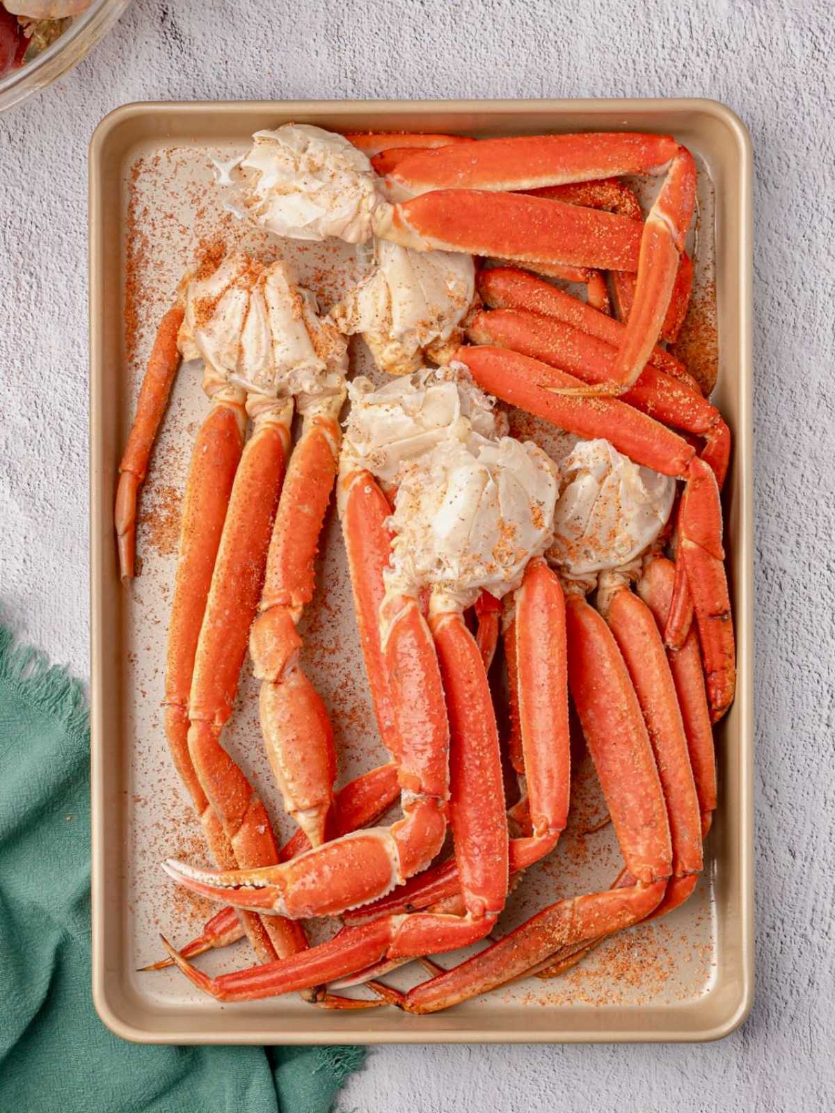 Cooked crab legs seasoned with spices on a baking sheet, placed on a light surface next to a green cloth.
