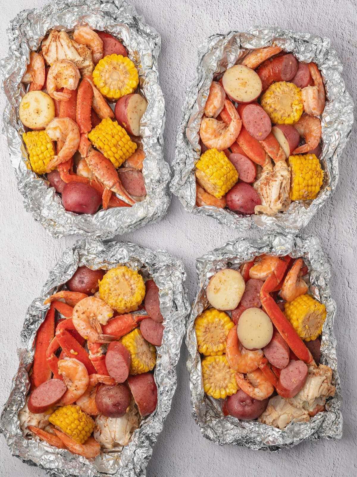 Four foil packets filled with shrimp, sausage, corn, potatoes, and crab legs on a light surface.